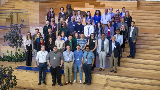 A large group of professionally dressed individuals poses for a group photo on tiered wooden seating inside a modern building with plants and a statue of a wildcat in the background. The group appears to be attending a conference or symposium, as many are wearing name badges and lanyards.
